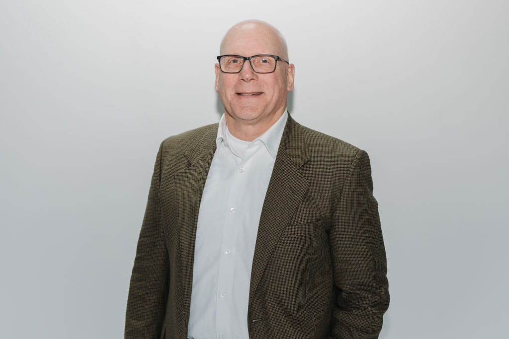 A bald man wearing glasses, a white shirt, and a brown checkered blazer stands against a plain light gray background, smiling slightly.
