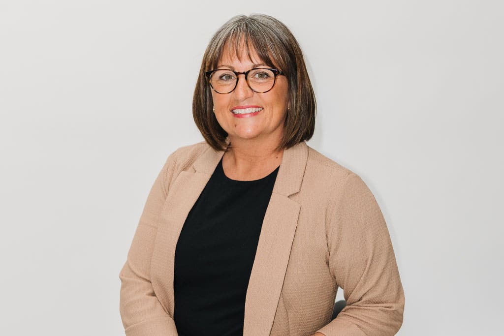 A woman with straight brown hair, wearing glasses, a beige blazer, and a black top, smiling in front of a plain white background.