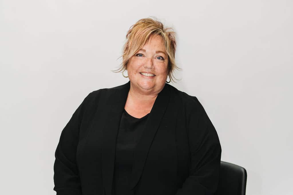 A woman with short, light brown hair, wearing a black top and black jacket, smiles while sitting on a chair against a plain white background.