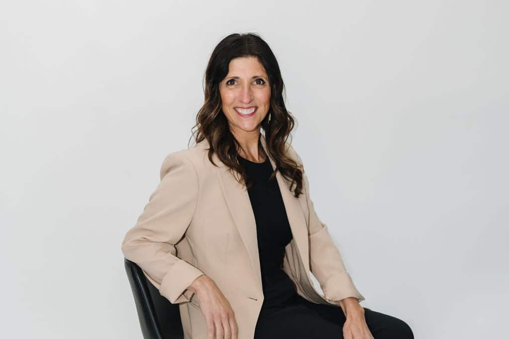 A woman with long brown hair, wearing a beige blazer and black top, sits on a black chair and smiles at the camera against a plain white background.