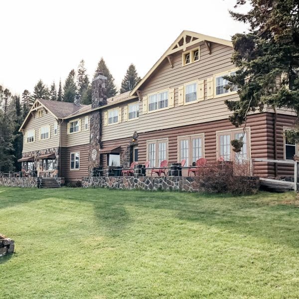 A large rustic lodge with log siding, stone chimney, and many windows sits surrounded by grass and tall pine trees. Red chairs line the patio, welcoming visitors much like local insurance agents ready to assist the community.