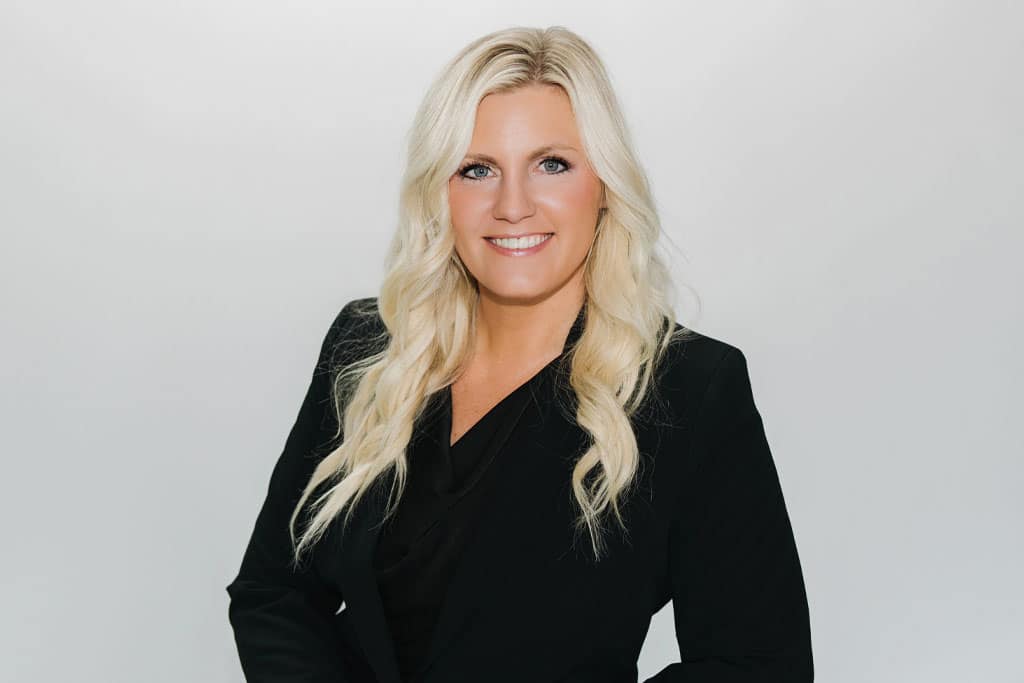 A woman with long, wavy blonde hair wearing a black blazer and black top, smiling at the camera against a plain, light background.