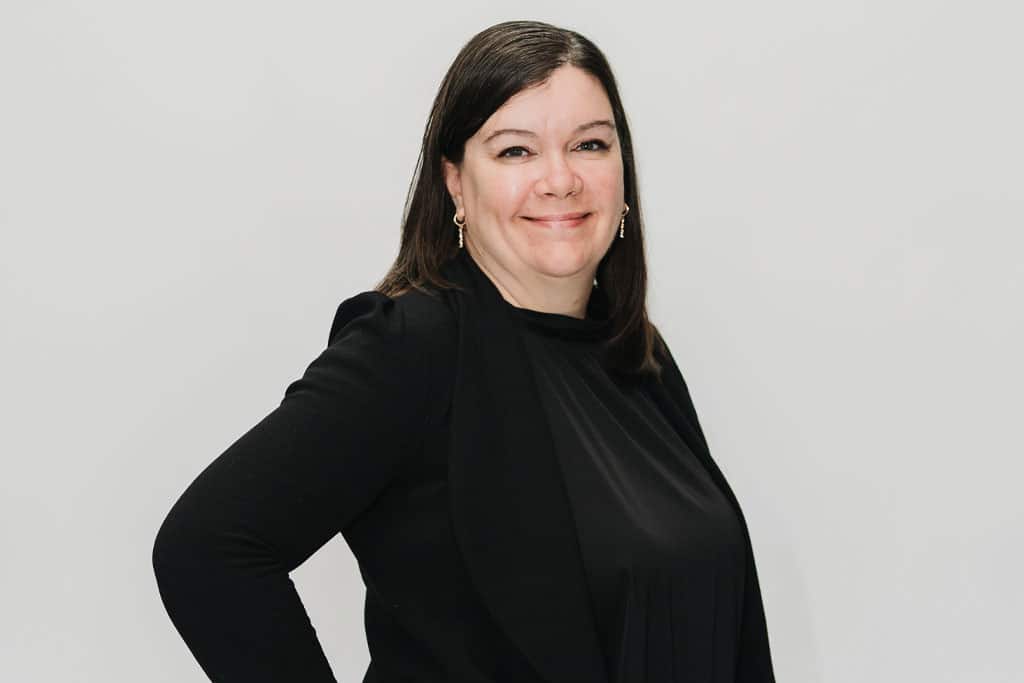 A woman with straight brown hair, wearing a black top and blazer, stands against a plain light background, smiling slightly with her hand on her hip.