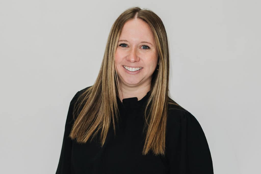 A woman with straight, light brown hair, wearing a black long-sleeve top, smiles at the camera against a plain light gray background.