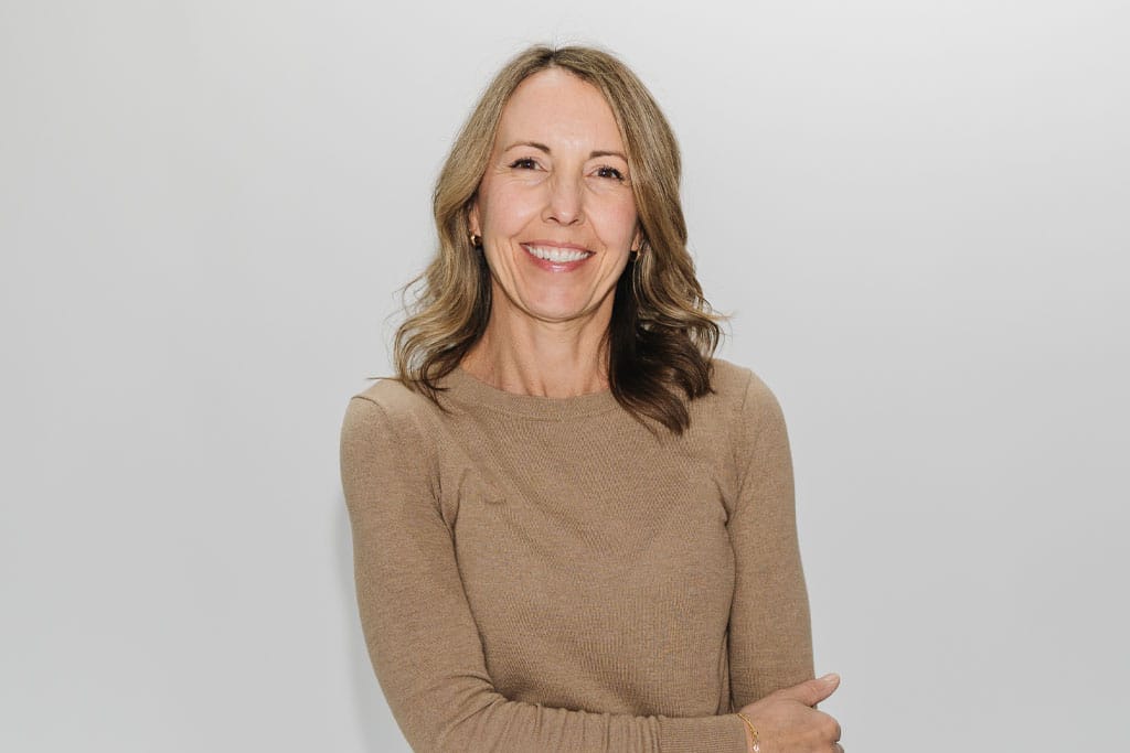 A woman with wavy light brown hair, wearing a tan long-sleeve top, smiles with her arms loosely crossed against a plain light gray background.