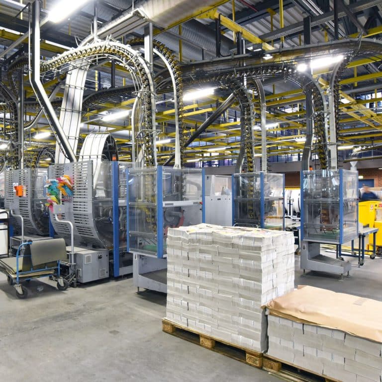 Stacks of packaged newspapers or magazines are on wooden pallets in a modern, automated printing facility—sometimes featuring ads from local insurance agents—amid conveyor belts, machines, and yellow metal framework overhead.