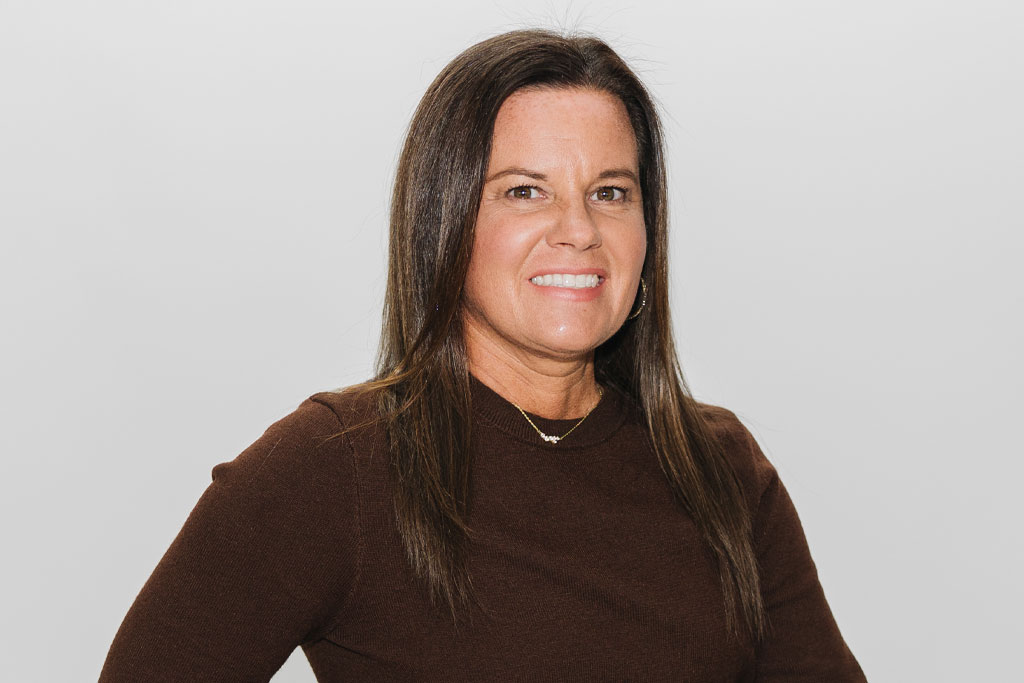 A woman with long straight brown hair, wearing a dark brown sweater and a necklace, smiles at the camera against a plain light gray background.