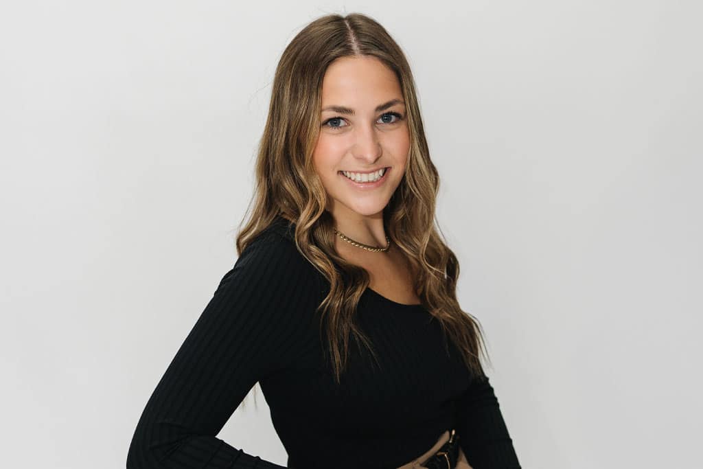 A young woman with long, wavy brown hair smiles at the camera. She is wearing a black, long-sleeve top and a gold necklace, standing against a plain white background.