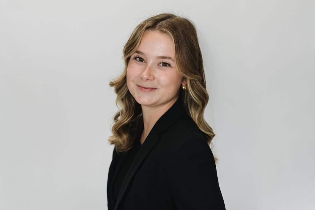 A young woman with wavy, shoulder-length blonde hair smiles slightly while wearing a black blazer. She is posed in front of a plain light-colored background.