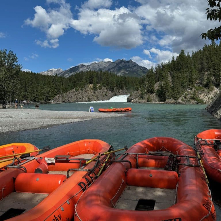 Bright orange rafts rest on the shore of a clear river surrounded by pine trees, with a waterfall and mountains in the background under a partly cloudy sky. Local insurance agents could help ensure your next adventure is worry-free.
