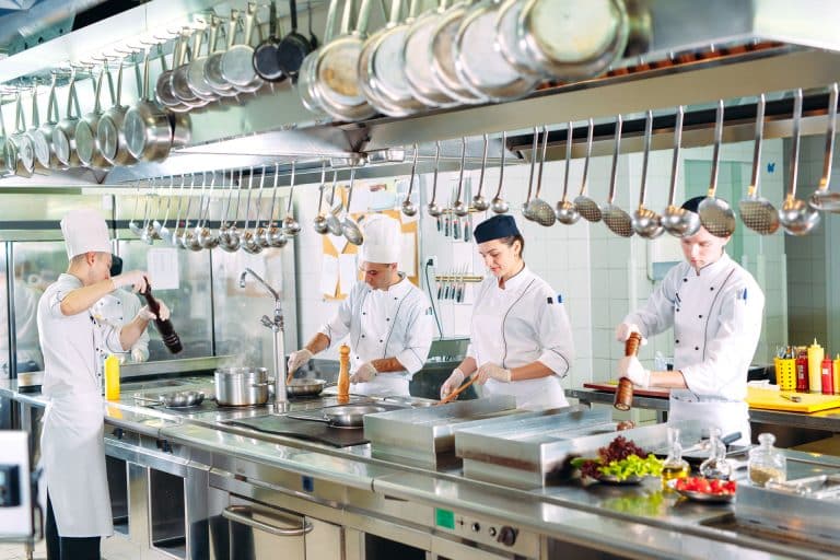 Four chefs in white uniforms work in a professional kitchen, preparing food at stainless steel counters. Like local insurance agents, they expertly handle each ingredient with care, ensuring everything comes together perfectly.