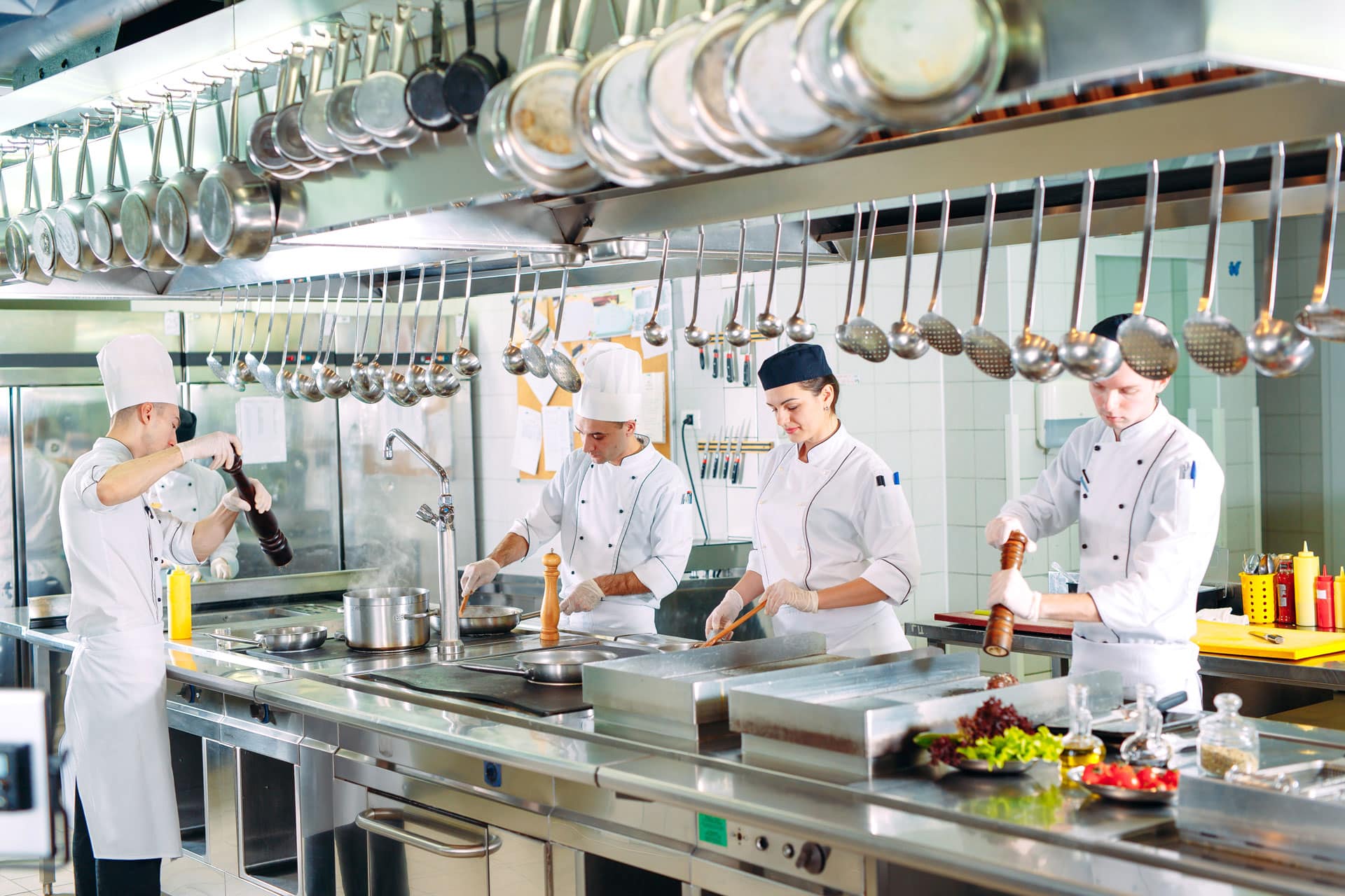Four chefs in white uniforms work in a professional kitchen, preparing food at stainless steel counters. Like local insurance agents, they expertly handle each ingredient with care, ensuring everything comes together perfectly.