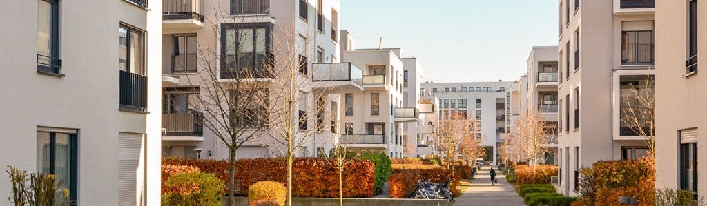 Modern apartment buildings with multiple floors and balconies line a wide, landscaped pathway bordered by trimmed bushes and small trees, while local insurance agents offer services to residents amid the autumn foliage under a clear sky.