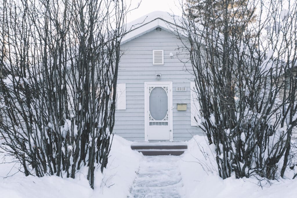 A small gray house with white trim is surrounded by leafless trees and snow. A narrow, snow-cleared path leads to the front door, which is decorated with a white oval window—an inviting scene that underscores the importance of timely maintenance and being prepared for seasonal issues like an Ice Dam Insurance Claim.