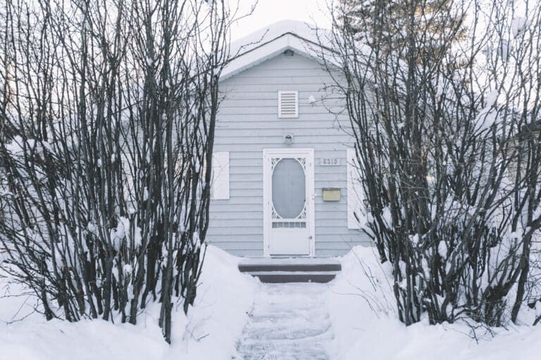 A small gray house with white trim is surrounded by leafless trees and snow. A narrow, snow-cleared path leads to the front door, which is decorated with a white oval window—an inviting scene that underscores the importance of timely maintenance and being prepared for seasonal issues like an Ice Dam Insurance Claim.