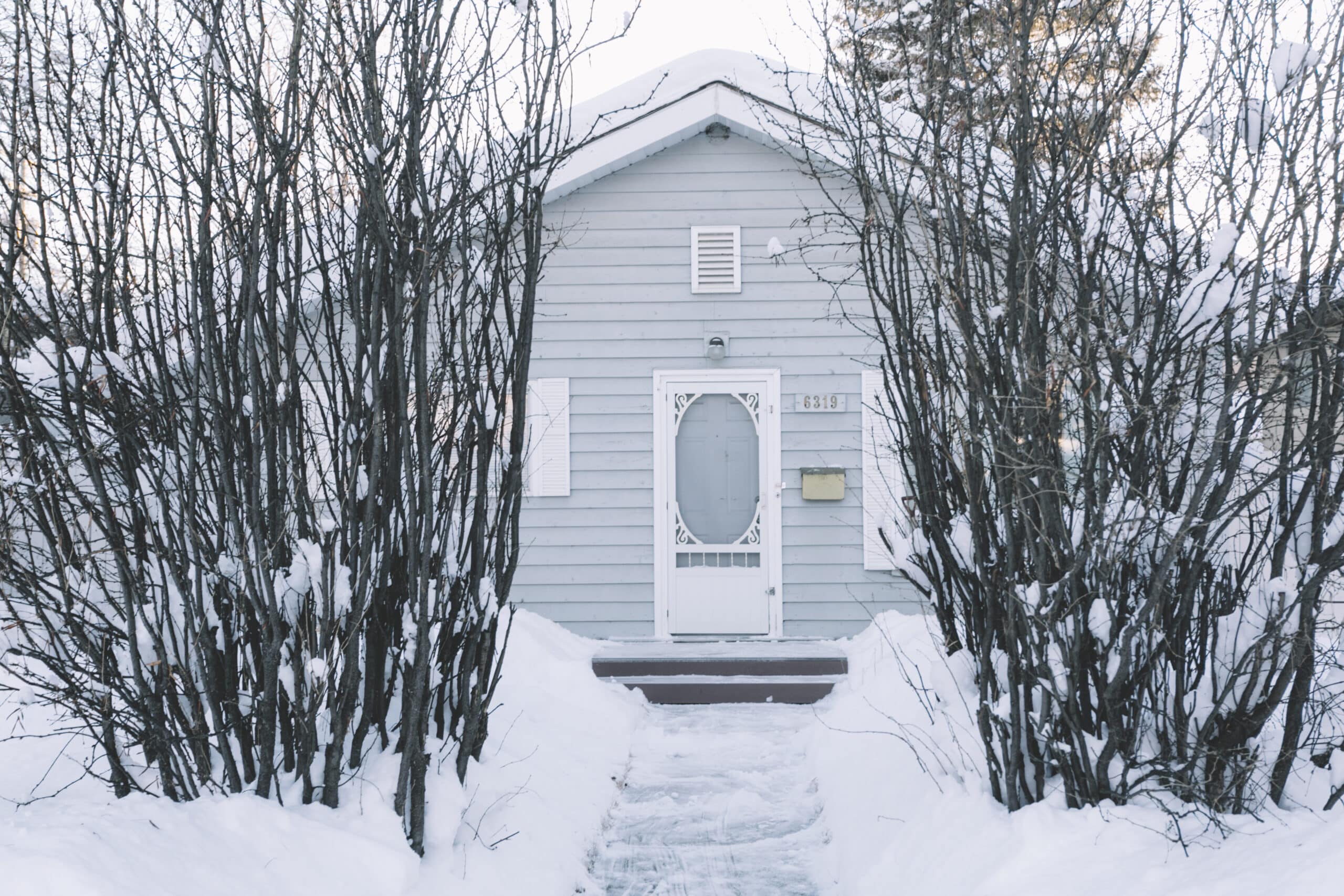 A small gray house with white trim is surrounded by leafless trees and snow. A narrow, snow-cleared path leads to the front door, which is decorated with a white oval window—an inviting scene that underscores the importance of timely maintenance and being prepared for seasonal issues like an Ice Dam Insurance Claim.
