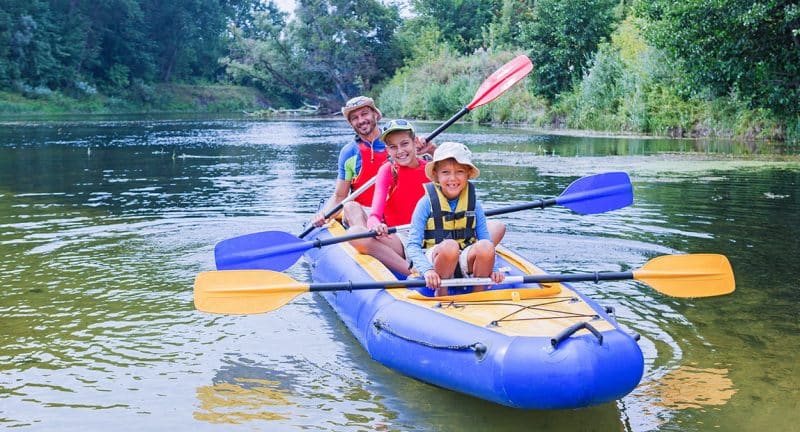 Three people, two children and one adult, smile and paddle together in a blue inflatable kayak on a calm river, enjoying the peace of nature—just the kind of moment local insurance agents help protect for families.